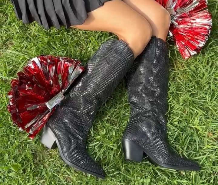 image of cheerleader wearing rhinestone embellished black western boots laying in the grass with two red pompoms 