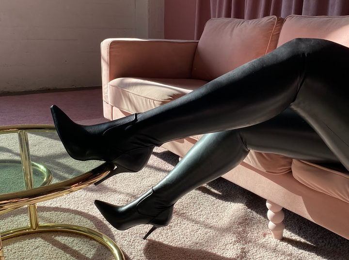 image of model wearing stretchy black thigh high stiletto boots sitting on a pink couch