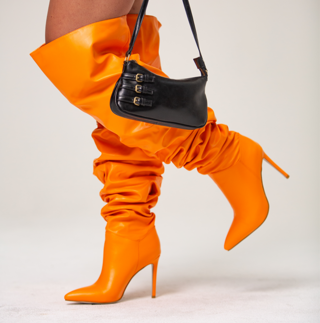 image of model posing while wearing bright orange knee high stiletto pointed toe boots with wide led scrunched boot shaft and holding black purse as an accessory