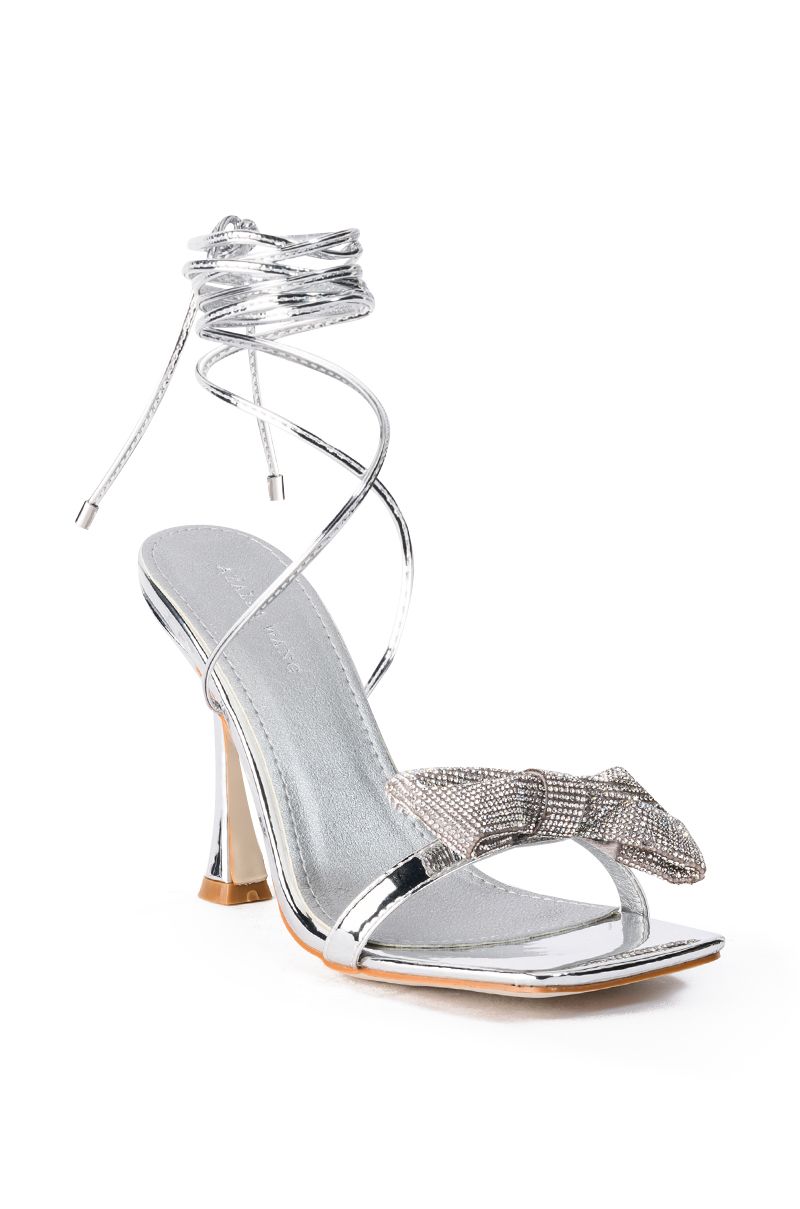 angled view of Silver stiletto heels with an open toe and square cut with silver wrap up cords and bow accent on the front strap